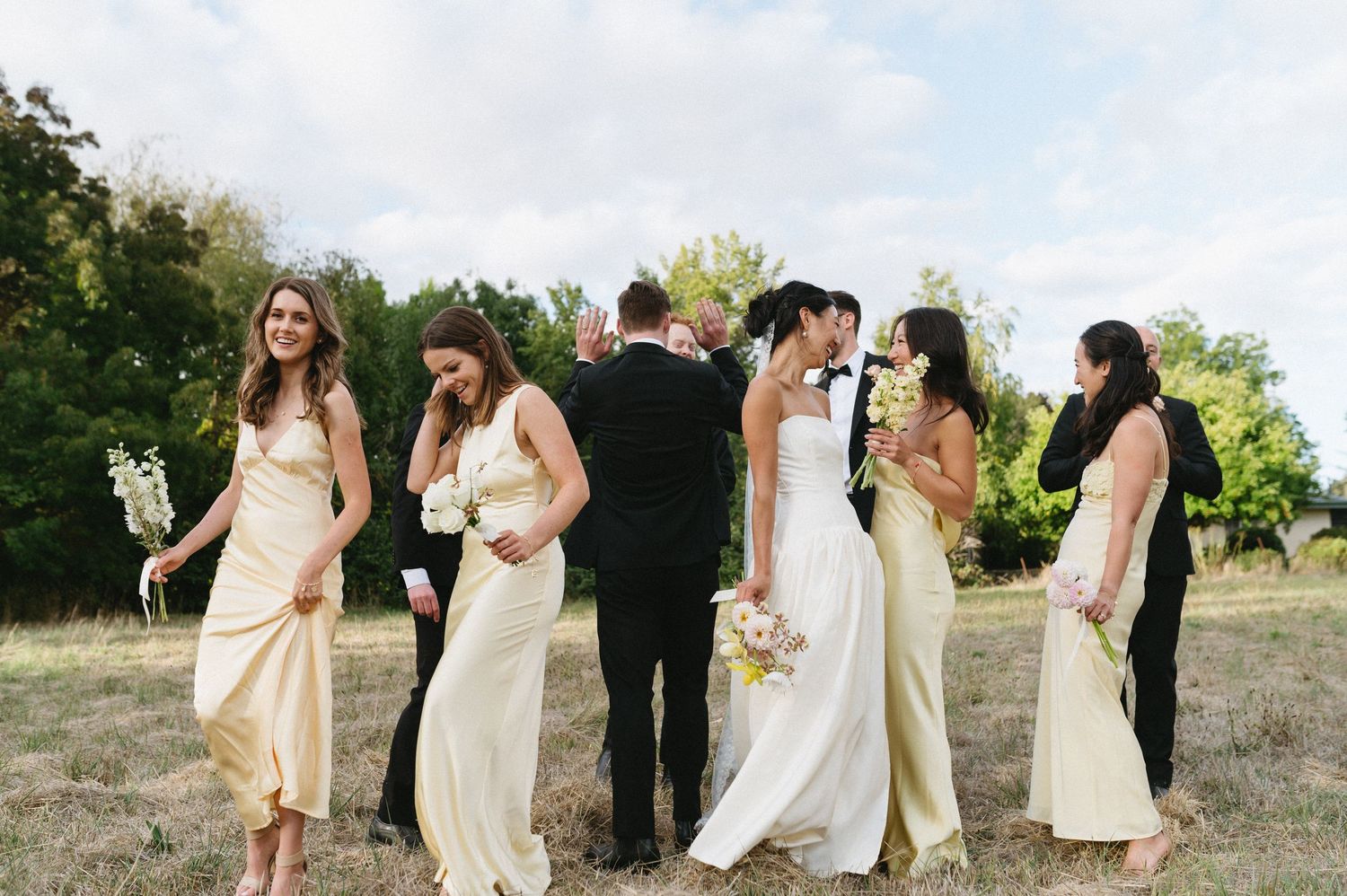 A bride in a modern white gown with asymmetrical neckline stands in sunlit garden setting.