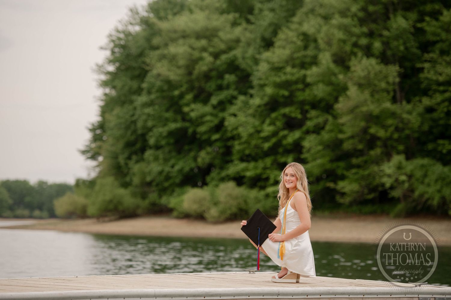 A person in a white dress crouches at the edge of a dock on a calm lake surrounded by green trees.