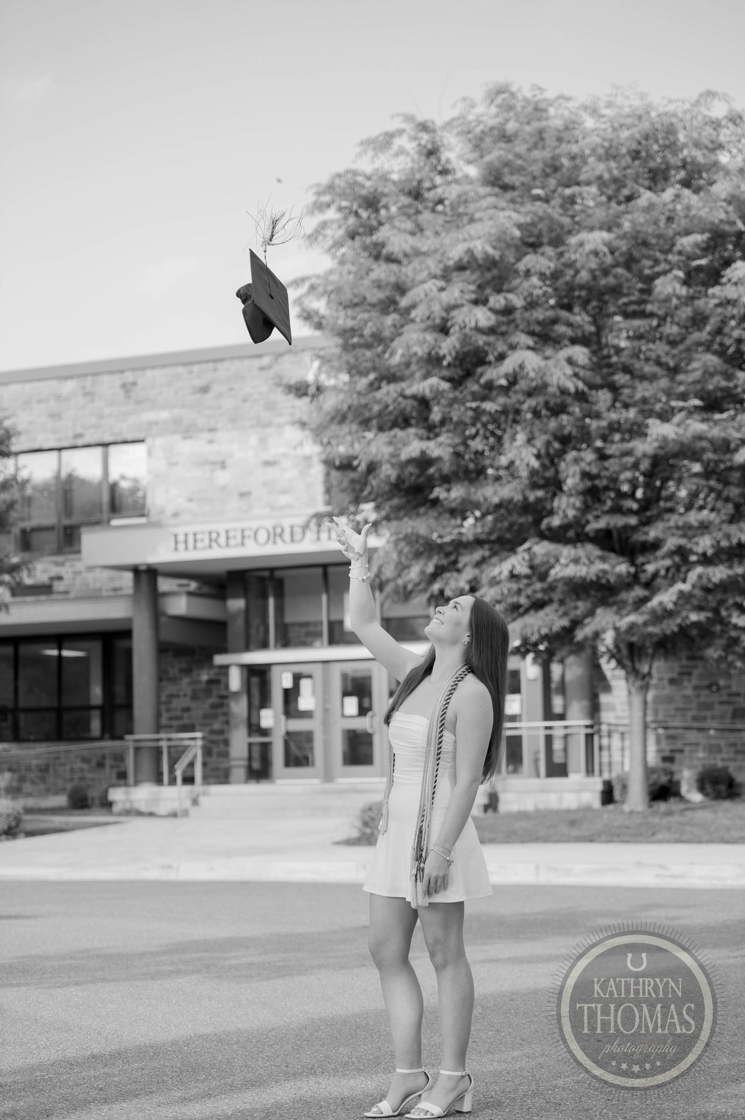 A graduation cap thrown in the air outside a school building with Hereford lettering visible in black and white.