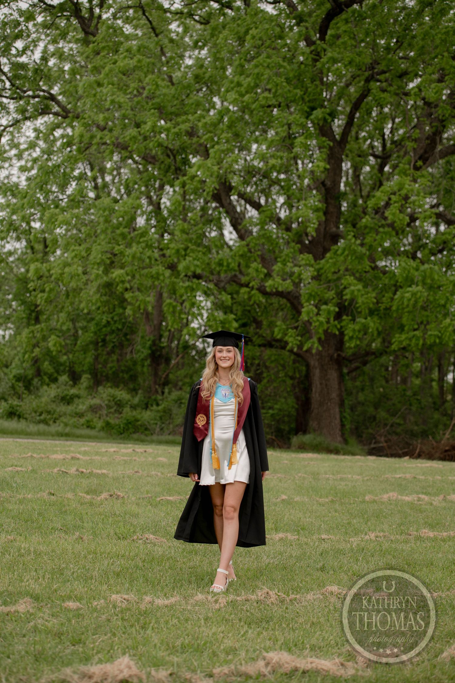 Graduate in cap and gown stands on grassy field with lush green trees in background during spring photoshoot.