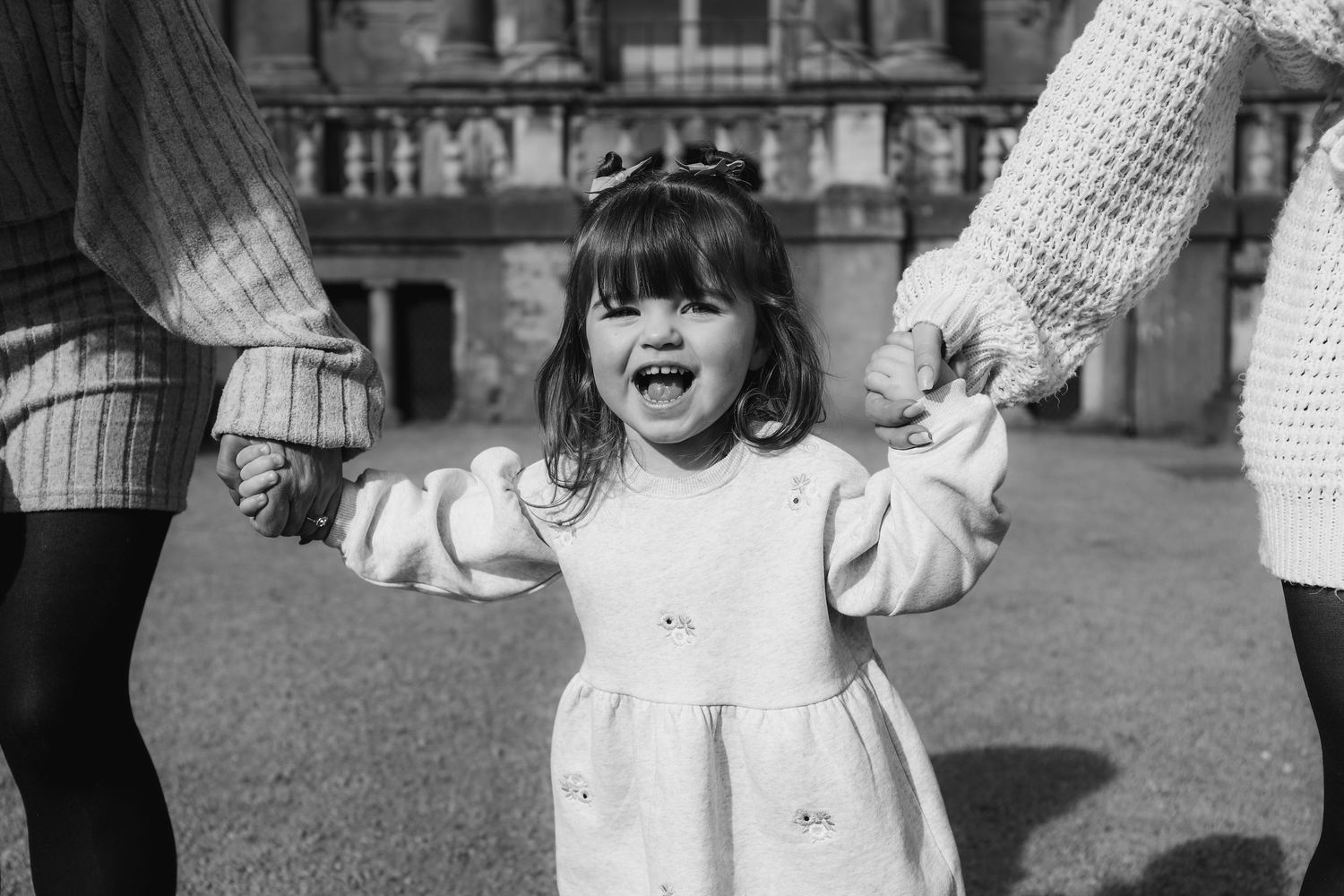 Black and white photo of a young child wearing a white dress holding hands with adults and smiling joyfully during Family shoot Nottingham