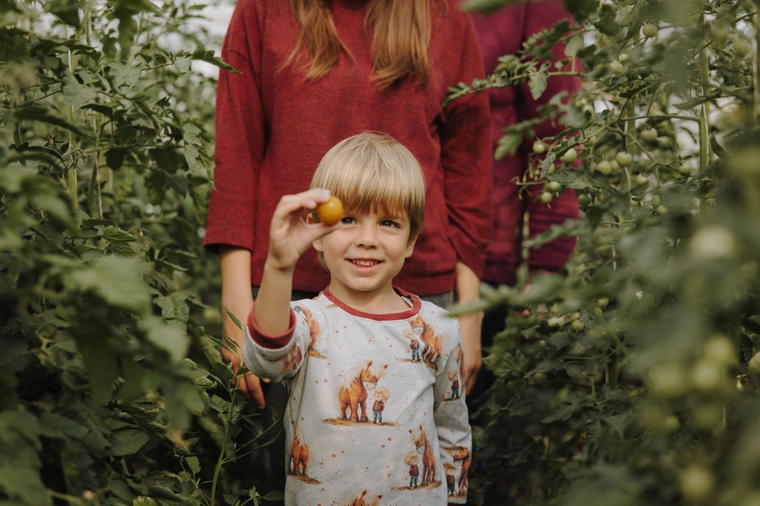 Child in red clothing holding produce near a greenhouse.