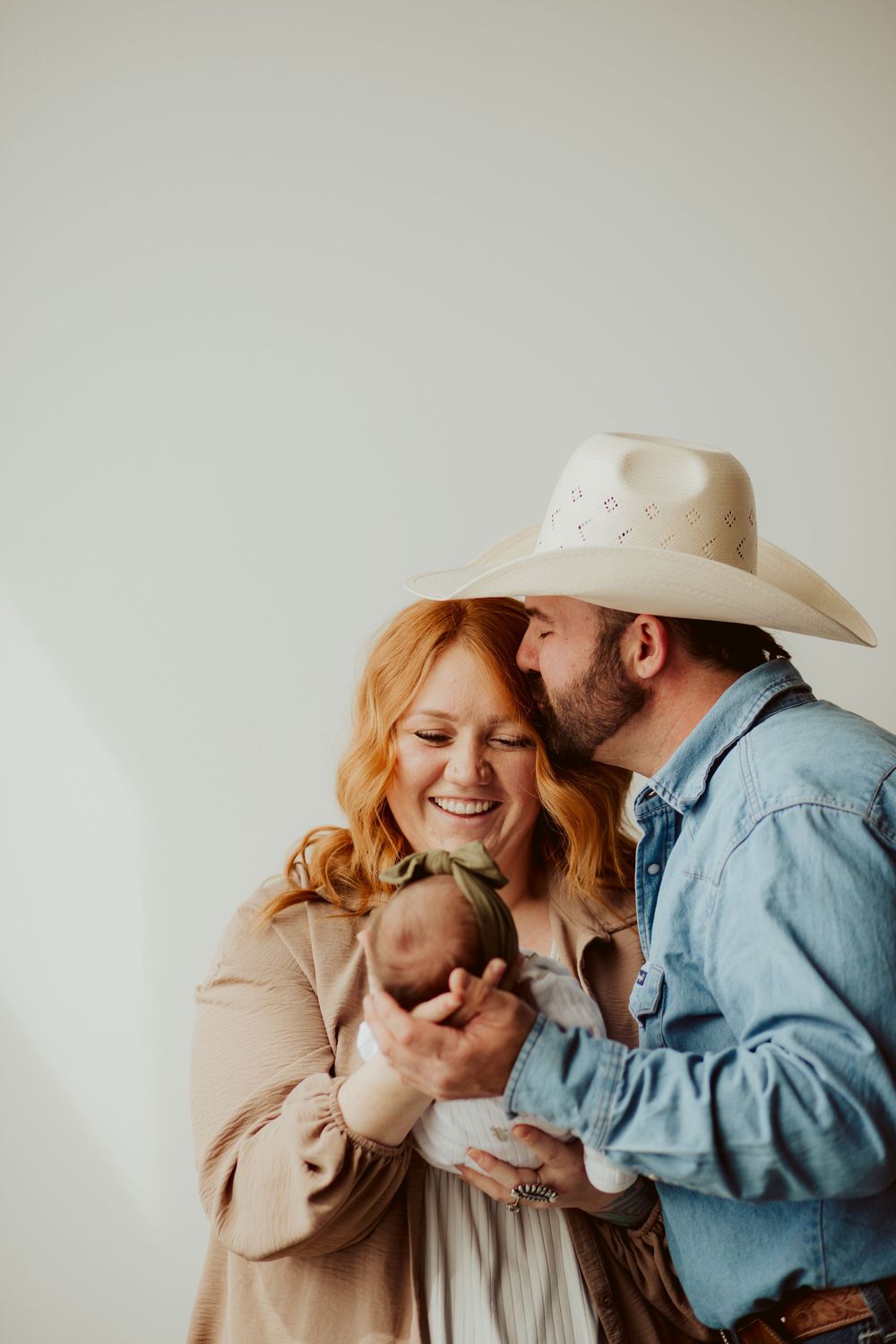 A couple in western attire shares a tender moment while holding their newborn baby against a light background.