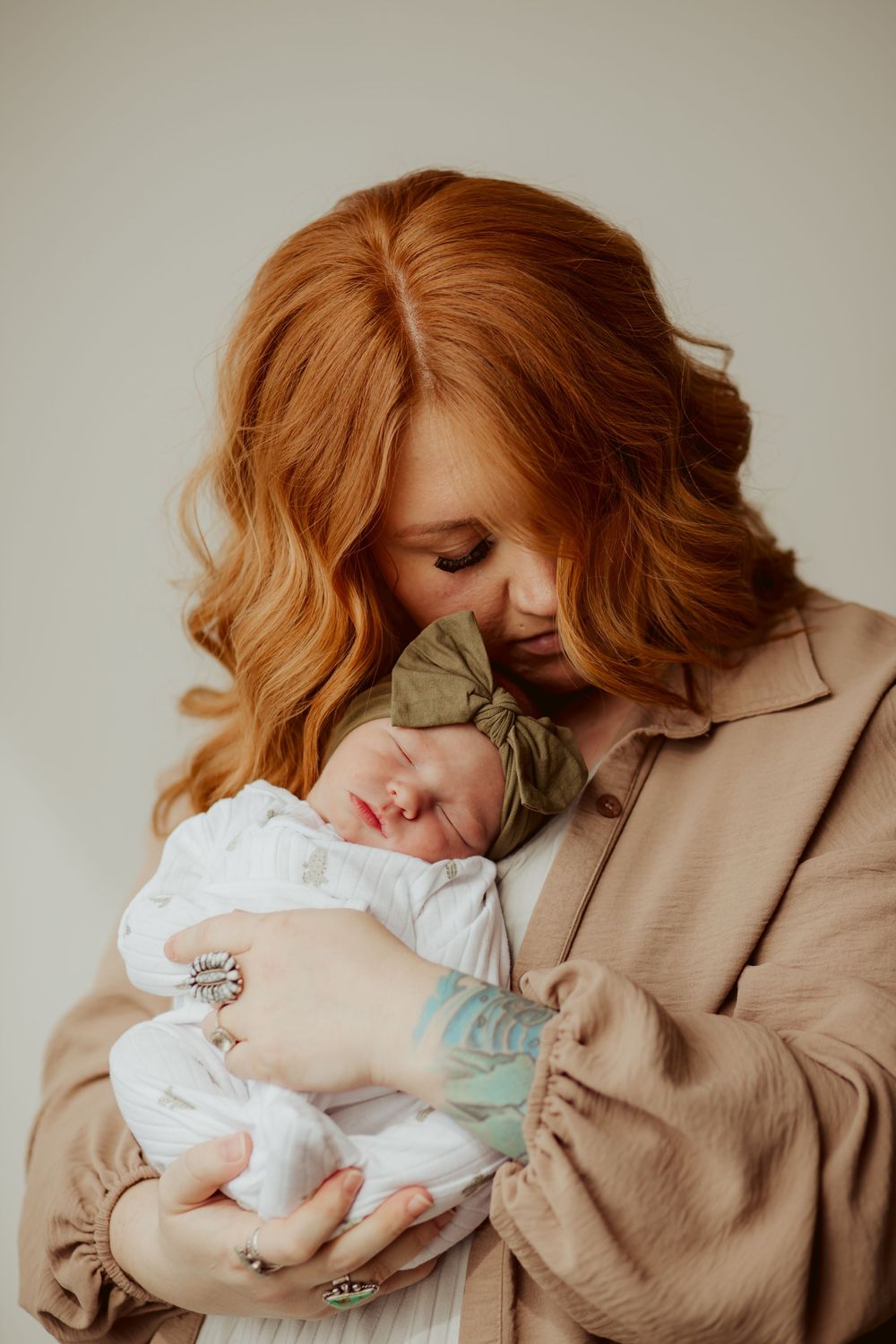 Woman in beige coat lovingly holds sleeping newborn baby wearing white outfit with green bow in warm neutral portrait.