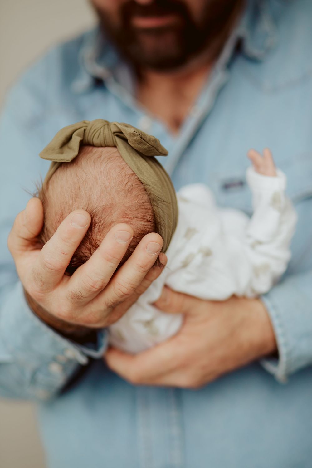 Someone in a denim shirt holds a newborn baby wearing white, with a beige bow headband visible on the infant's head.