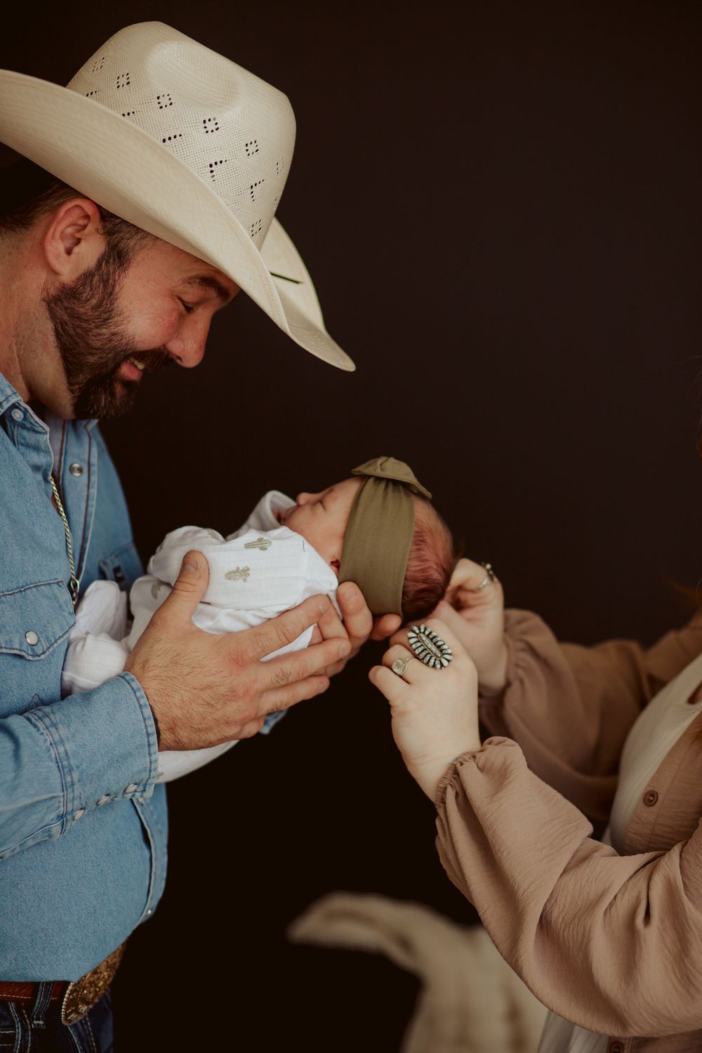 A person wearing a cowboy hat and denim shirt tenderly holds a newborn baby while another person watches.