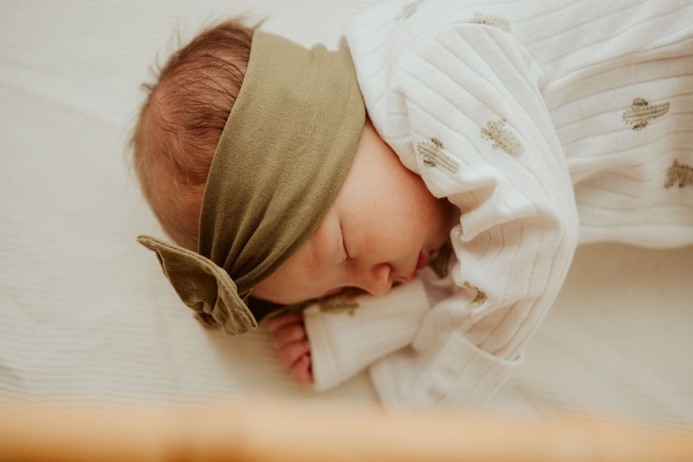 A sleeping baby wearing a sage green headband and white clothing lies peacefully on light beige fabric.