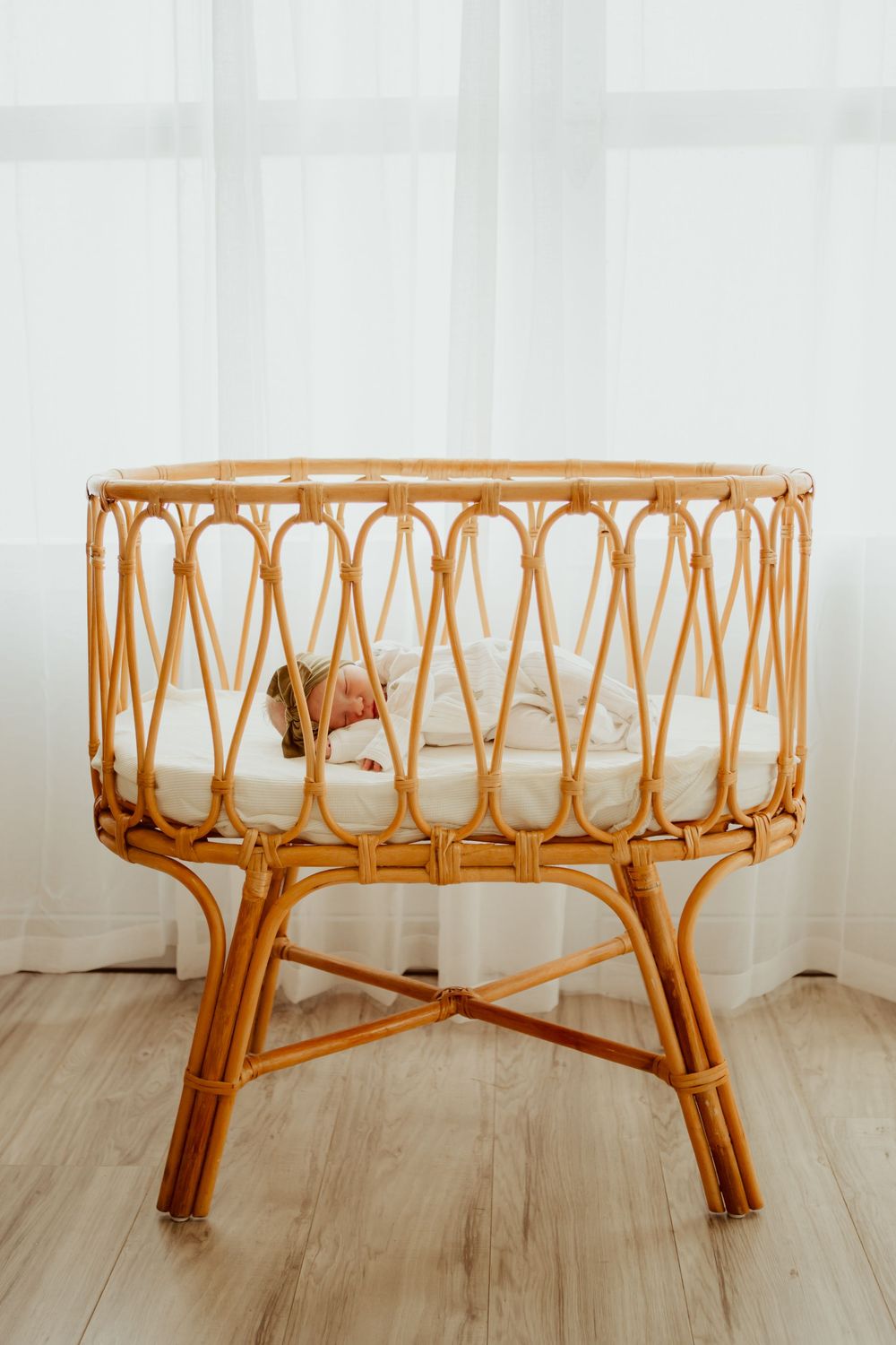 Vintage rattan baby bassinet crib with white cushioning stands elegantly on wooden flooring against light curtains.