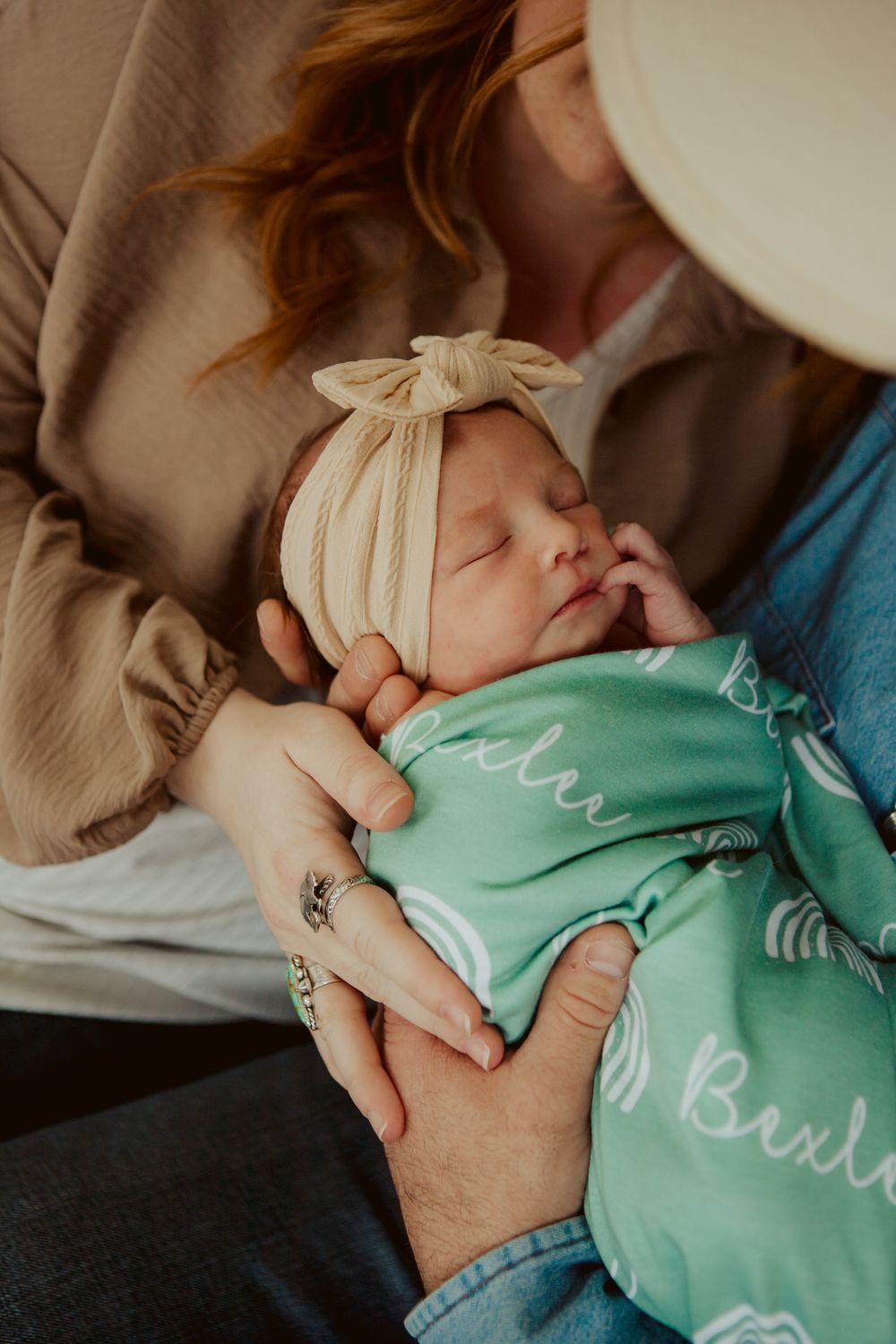 A sleeping newborn in a cream headbow wrap and mint green swaddle is cradled gently in caring arms.