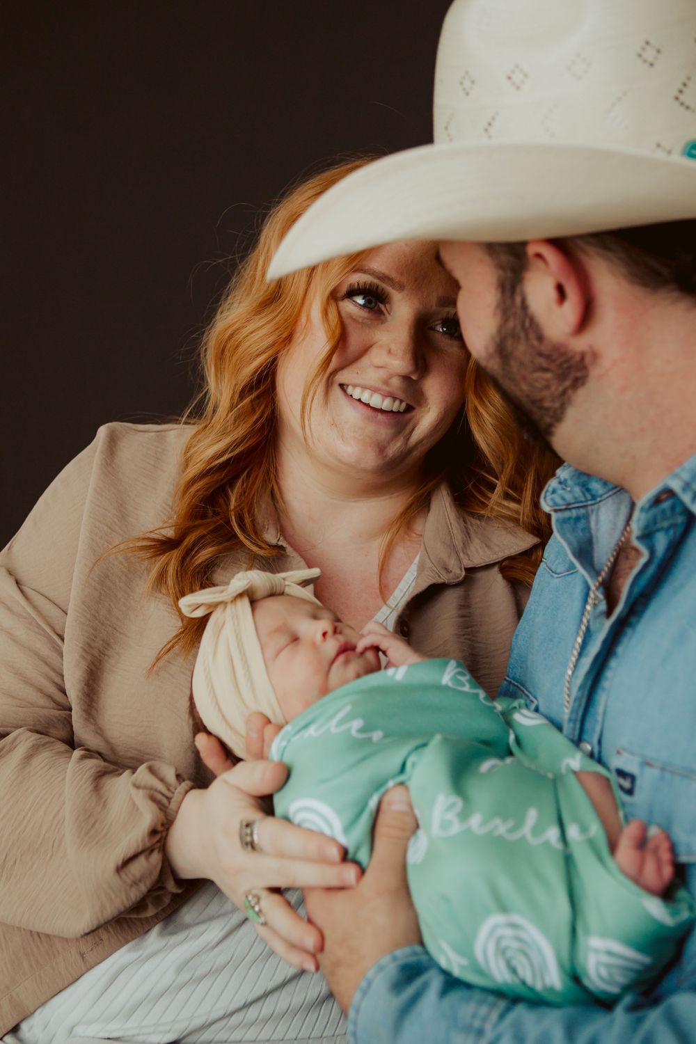 A couple smiles while holding their newborn baby wrapped in a mint green swaddle blanket.