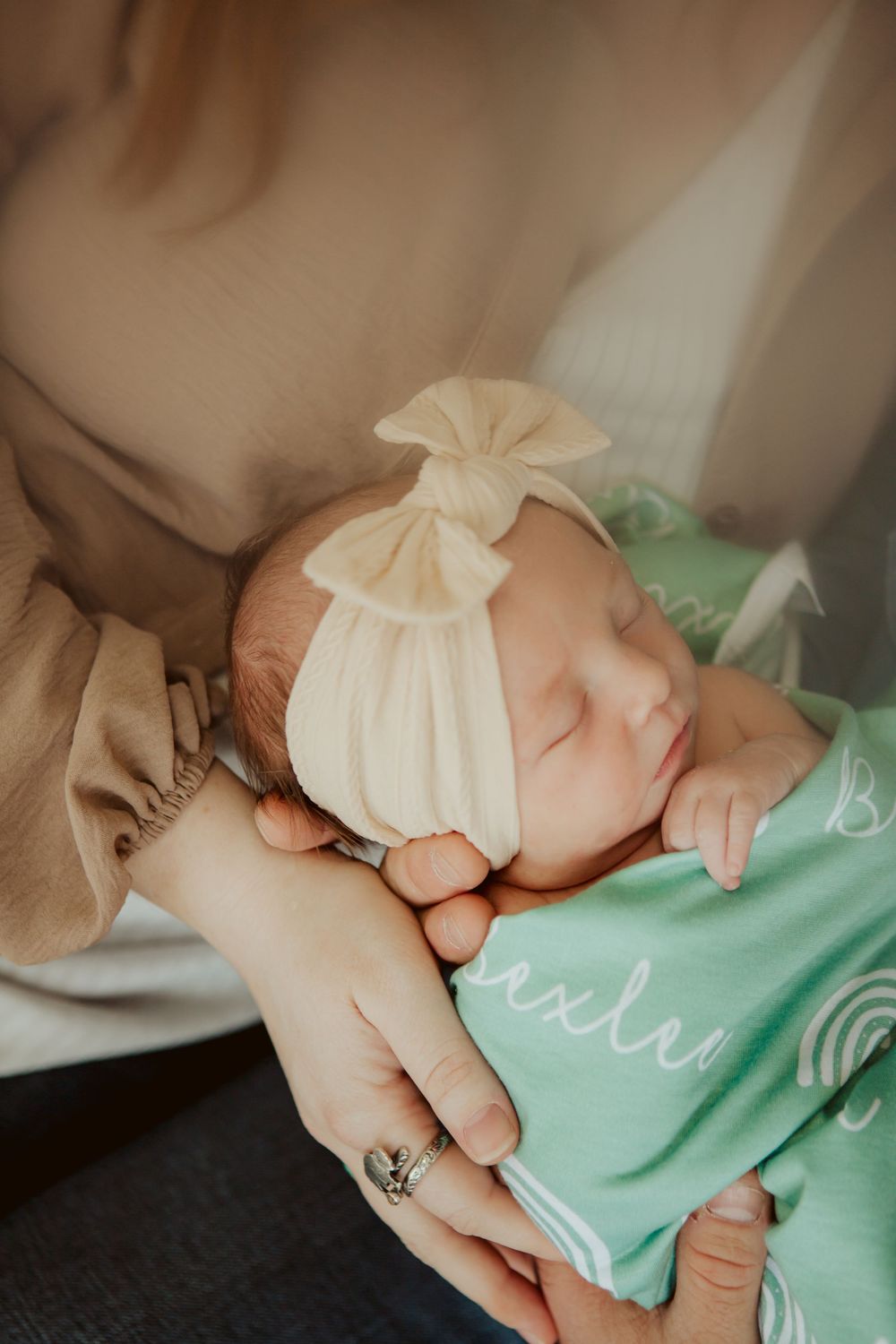 A sleeping newborn wearing a cream bow headband and mint green outfit rests peacefully in caring arms.