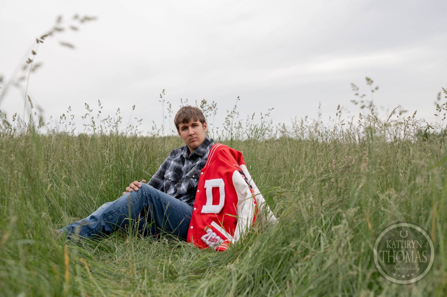 Student wearing high school letterman jacket sitting in tall grass field on cloudy day.