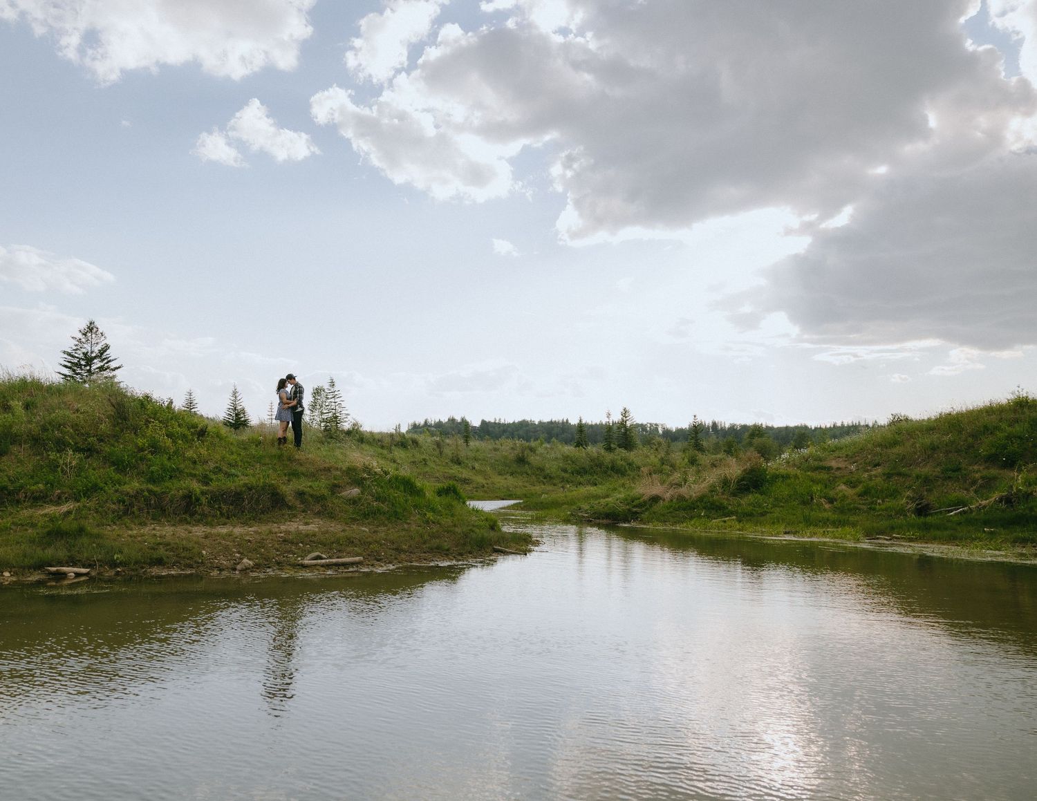 A lone figure stands on a grassy hilltop overlooking a peaceful meandering river under cloudy skies.