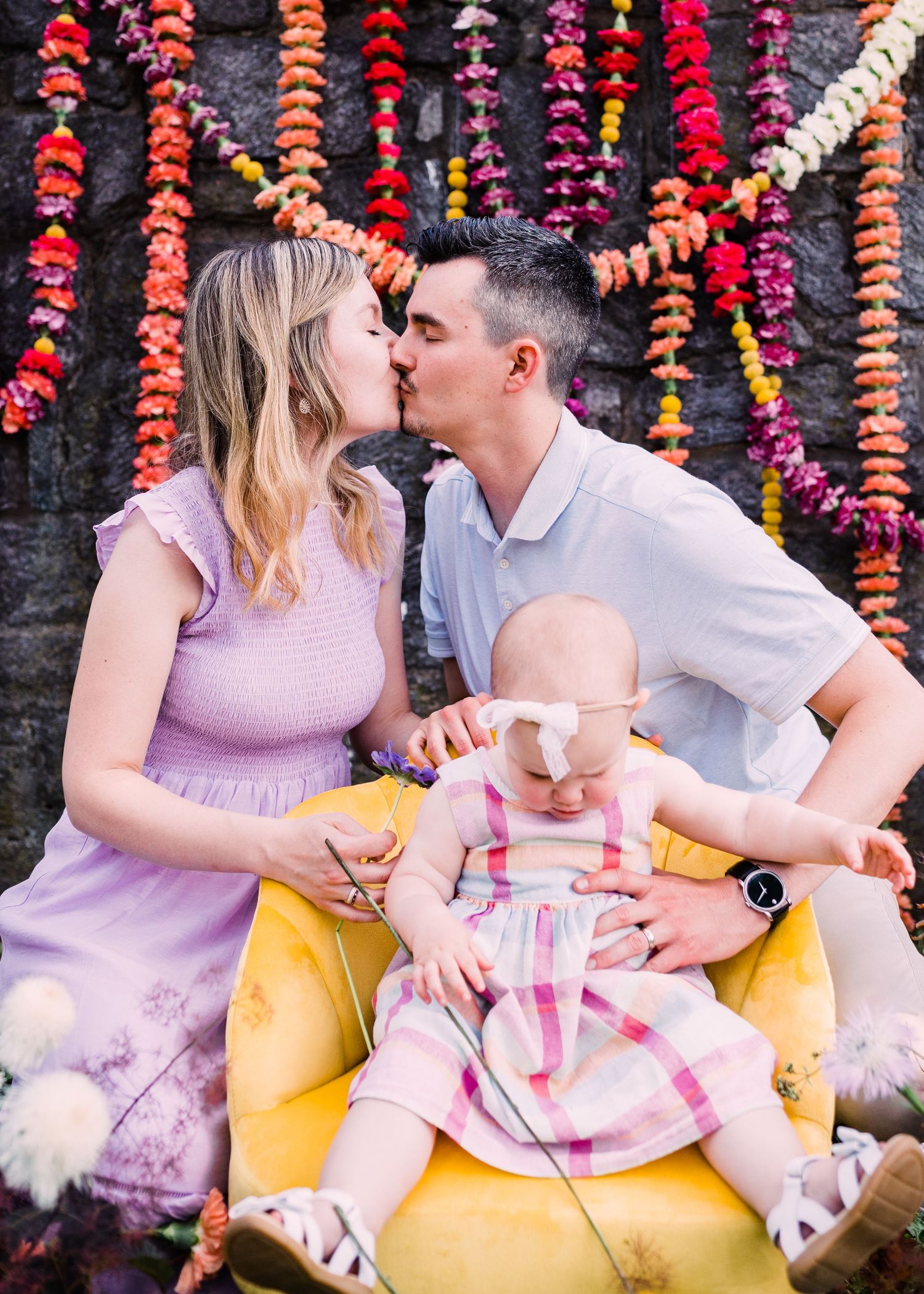 Two people sharing a romantic kiss in front of a colorful floral backdrop while sitting with a baby on a yellow chair.