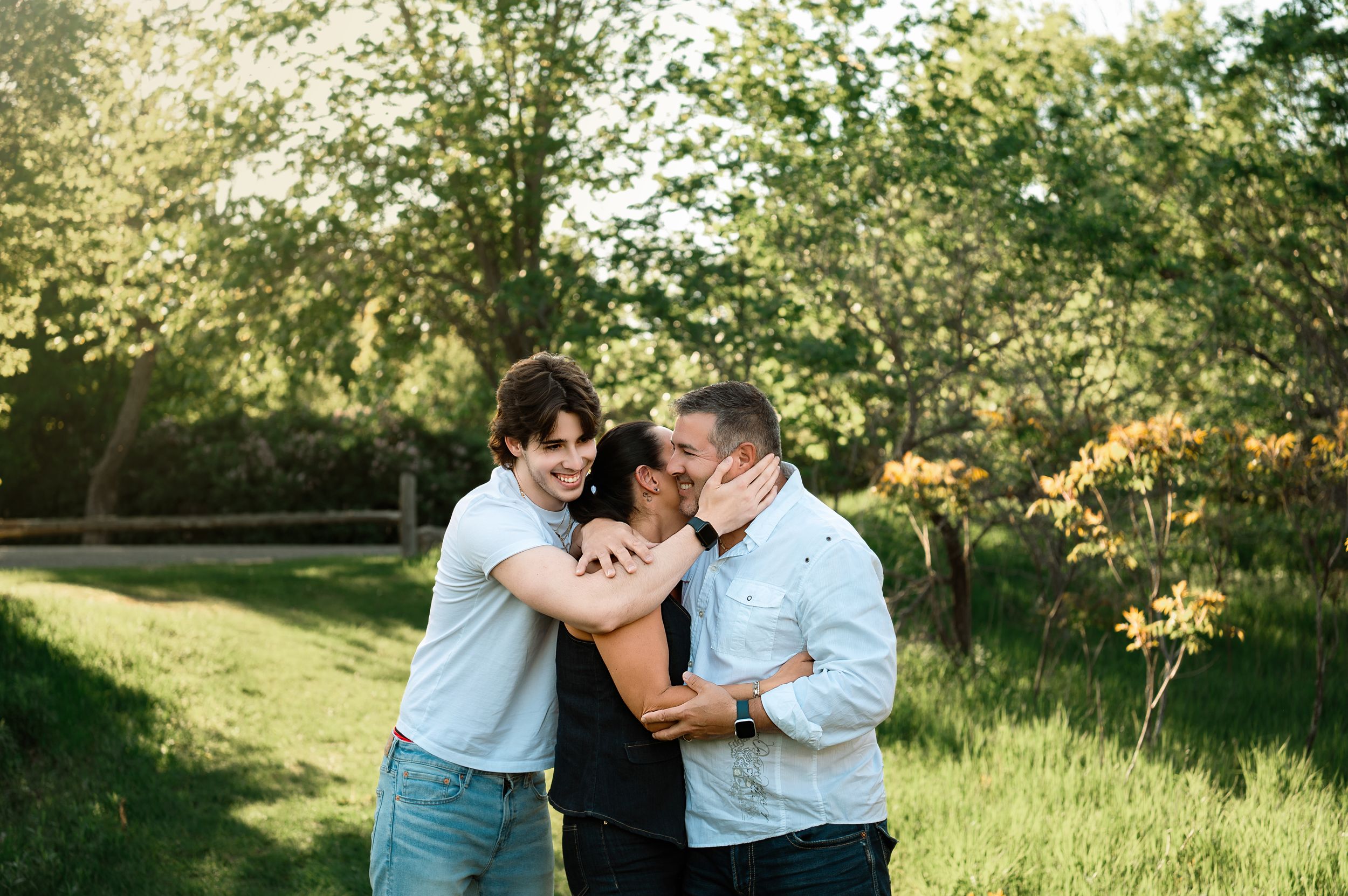 A joyful outdoor scene with three people embracing in a sun-dappled meadow surrounded by trees and green foliage.