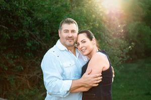 A couple embraces outdoors in a garden setting with warm sunlight glowing through trees in the background.