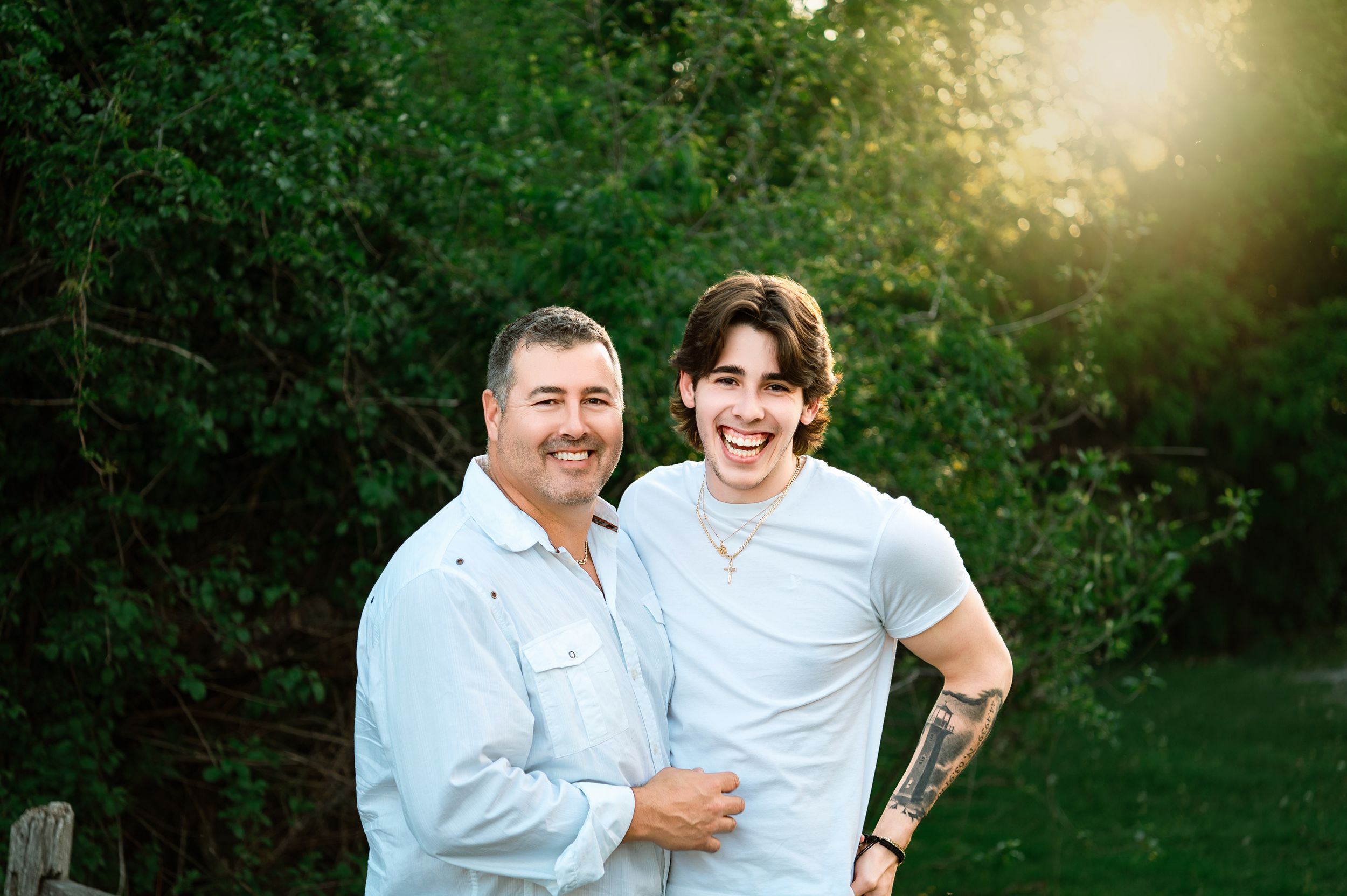 Two people wearing light colored shirts share a warm embrace while standing in front of lush green foliage in sunlight.