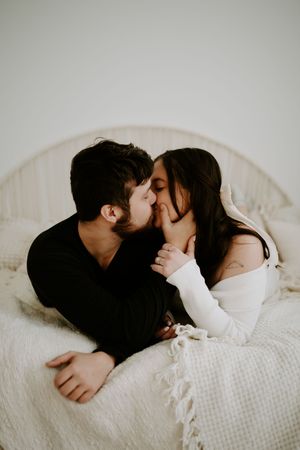 Romantic couple sharing tender moments on white bedding during intimate photoshoot.
