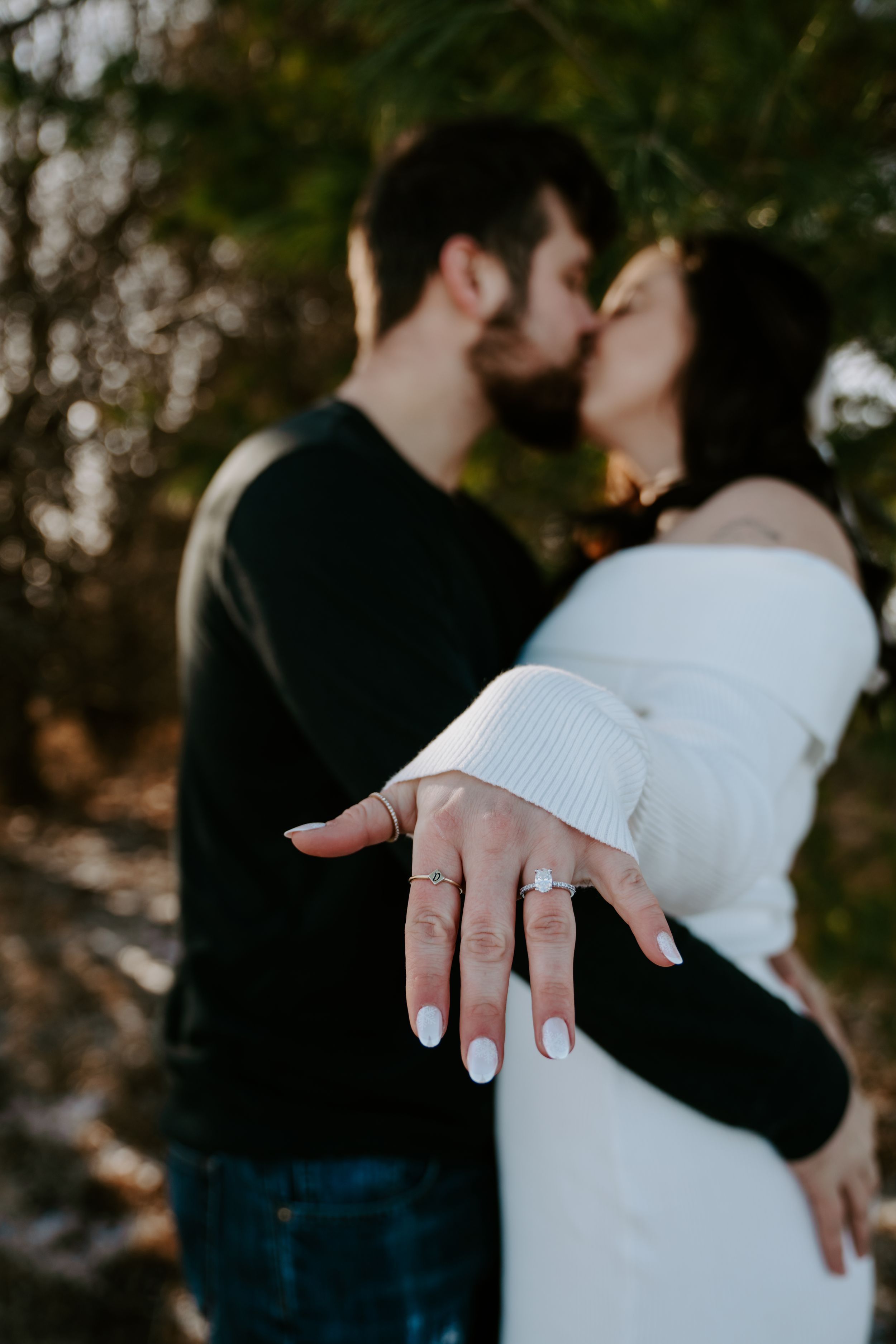 Engagement ring visible on hand while couple embraces in outdoor setting.