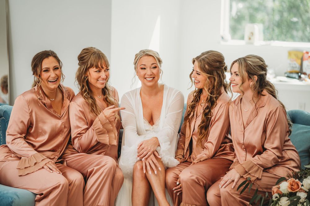 Bridal party in matching dusty rose robes shares a candid moment on blue couch.
