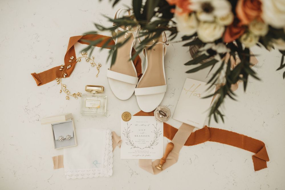 Wedding details flatlay with white shoes, perfume, rings and floral arrangement.