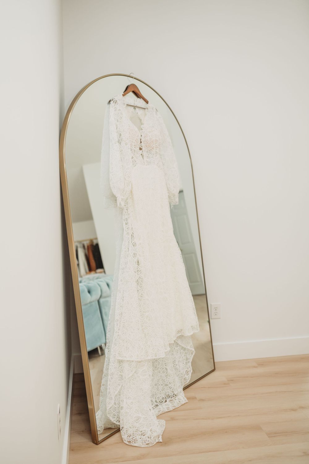 Elegant lace wedding dress hangs on display in arched full-length mirror against white wall.