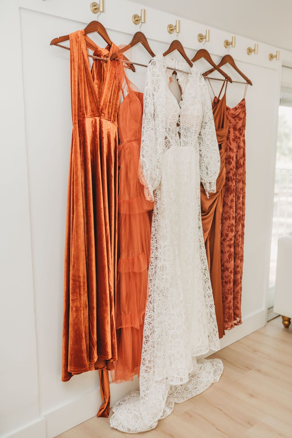 Wedding dress and bridesmaid dresses in rust and coral tones hang on wooden hangers.