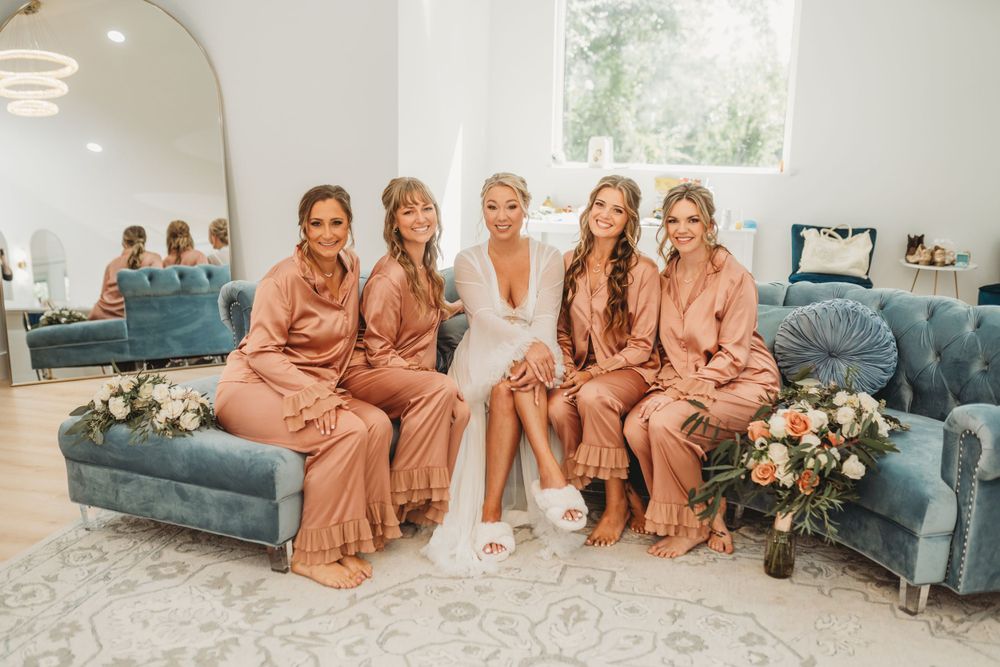 Bridal party in matching pink satin robes sits together on blue velvet couch.