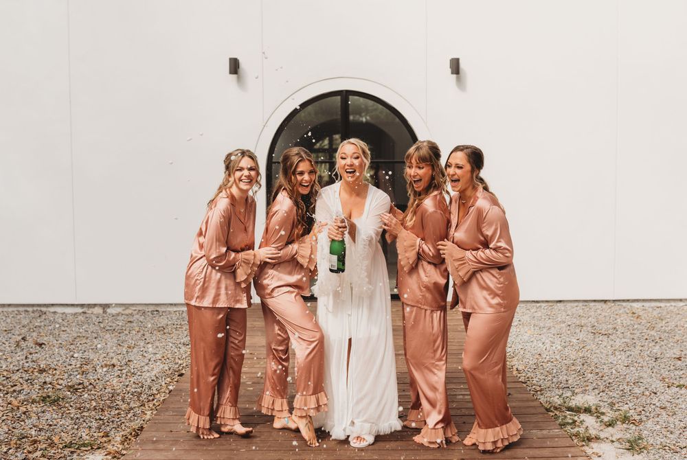 Wedding party poses together outdoors in coordinated rose gold satin robes against white wall.