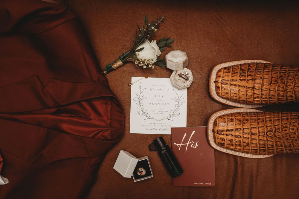 Wedding details including brown dress shoes, rings, and stationery arranged on burgundy fabric.