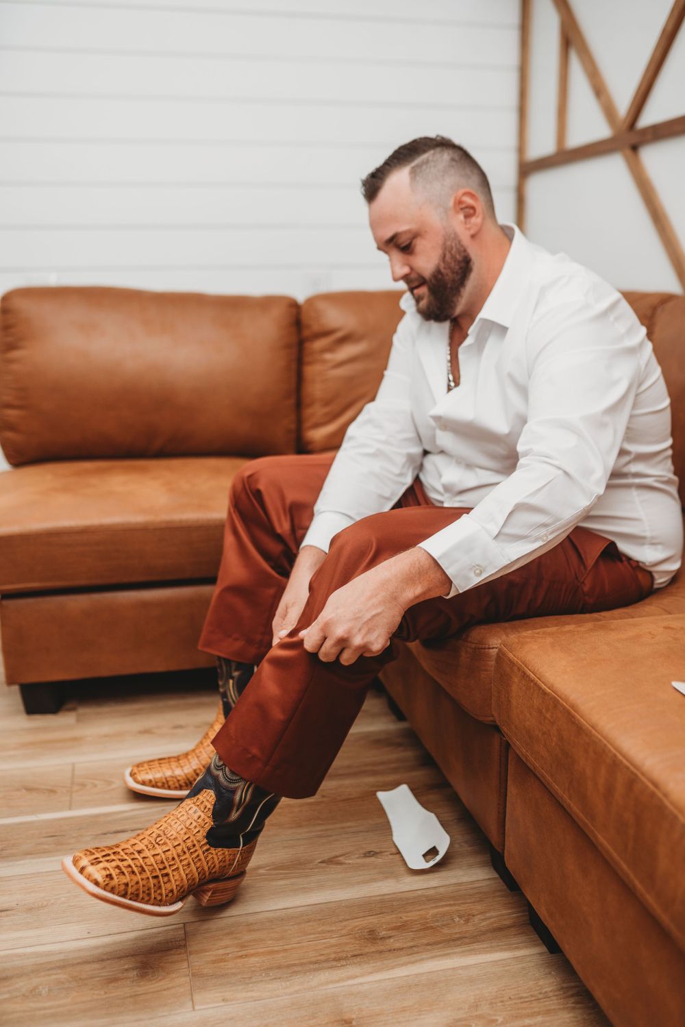 Person in white shirt and brown pants sits on leather couch while putting on shoes.