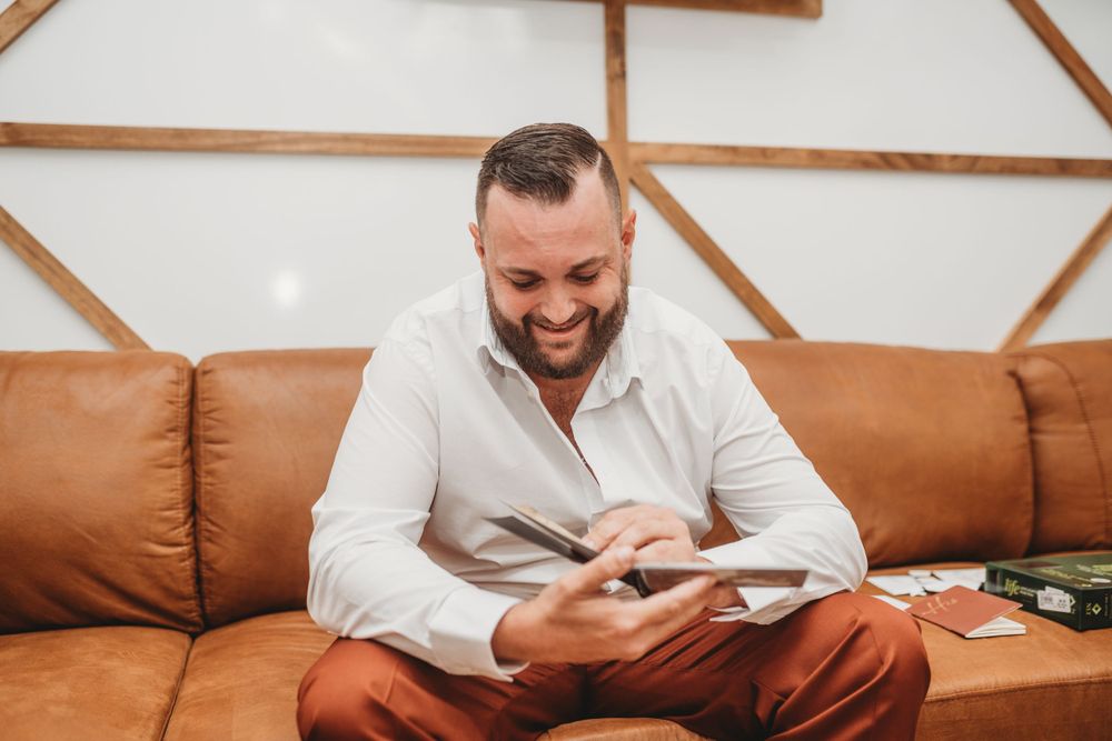 Person in white shirt and rust pants smiles while reading on brown leather couch.
