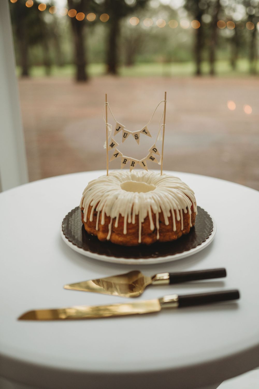 A bundt cake with white drizzle icing and banner topper displayed with gold serving utensils.