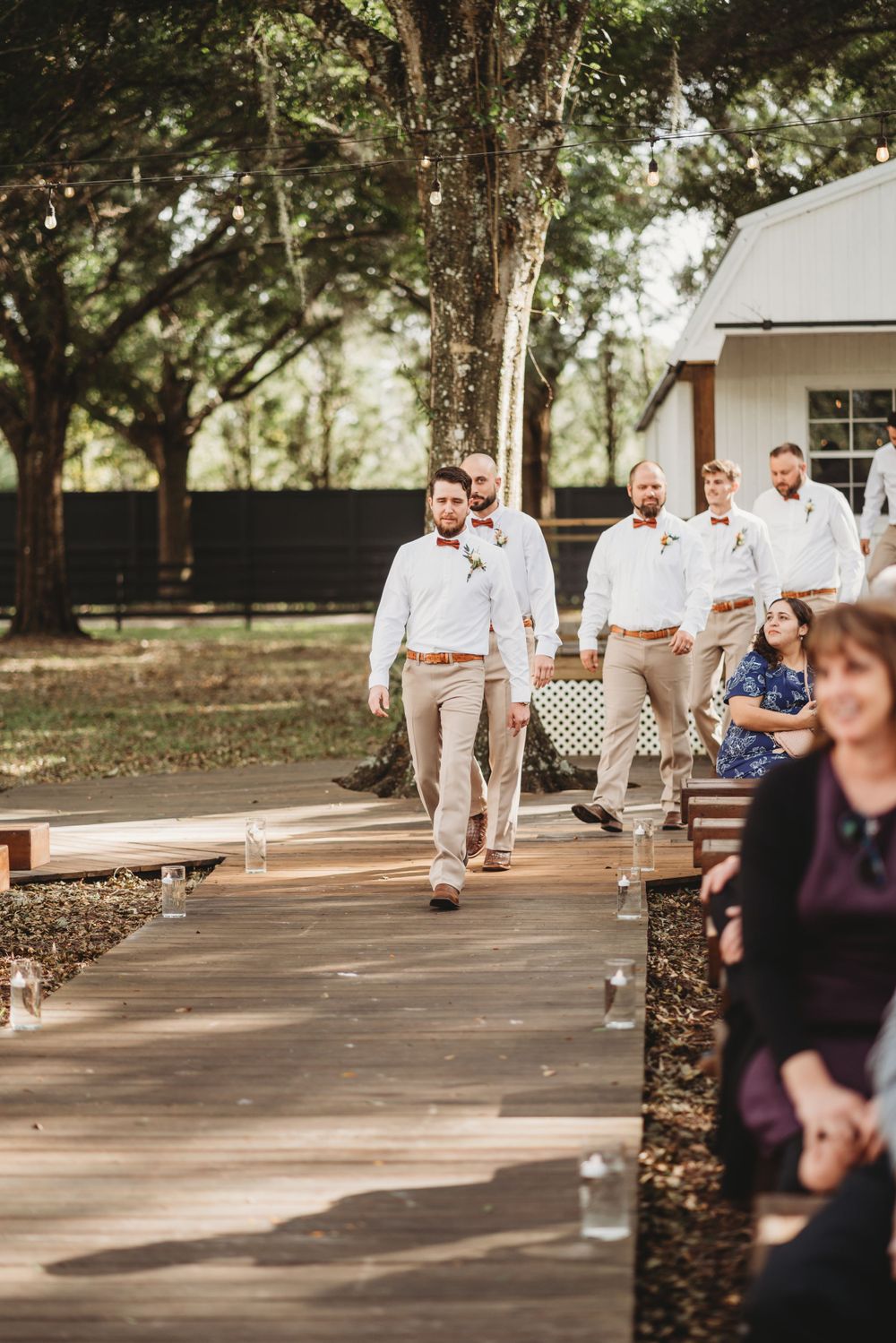 Groomsmen in light colored attire walk down an outdoor wooden pathway during a wedding ceremony.