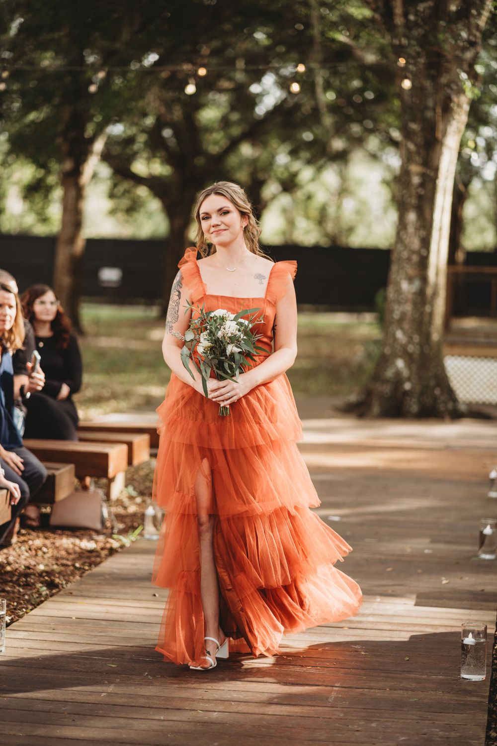 A bridesmaid walks down an outdoor aisle wearing a flowing coral tiered dress while carrying a bouquet.