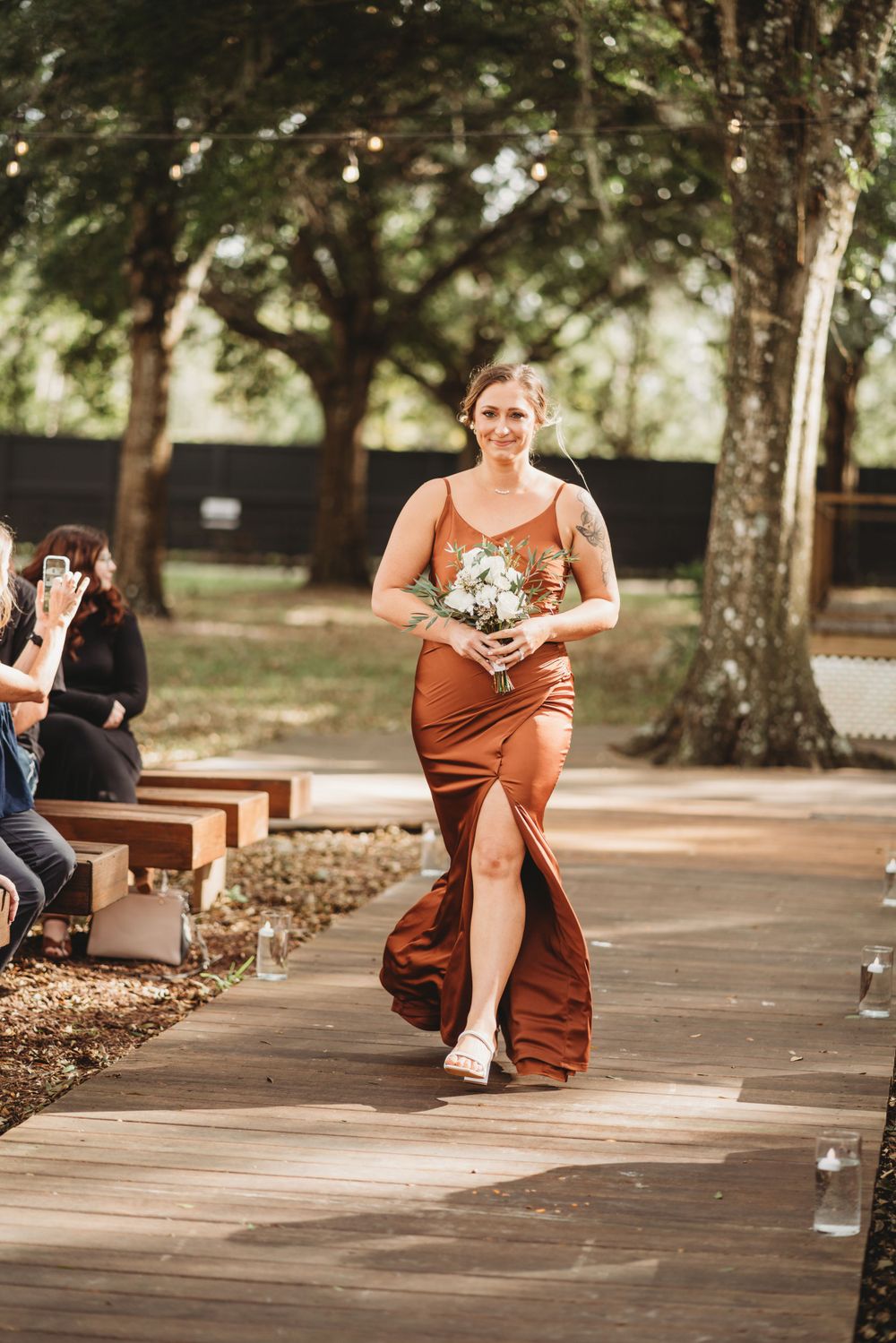 A bridesmaid in a rust-colored dress walks down an outdoor ceremony aisle carrying a small bouquet.