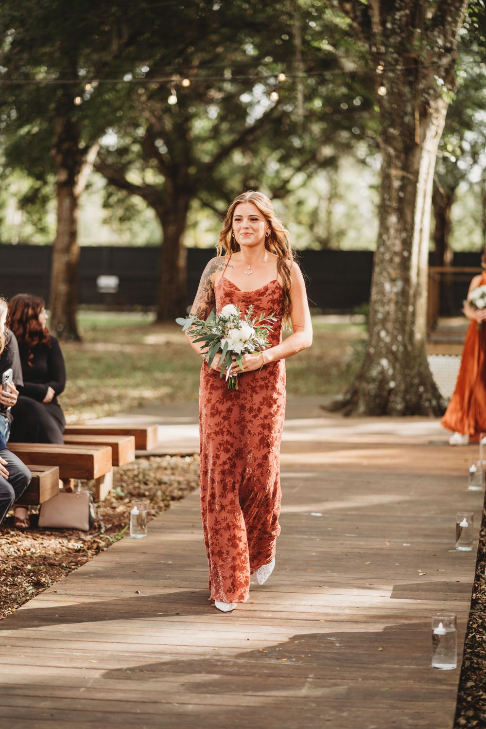 A bridesmaid walks down a wooden pathway carrying flowers and wearing a coral-colored dress.