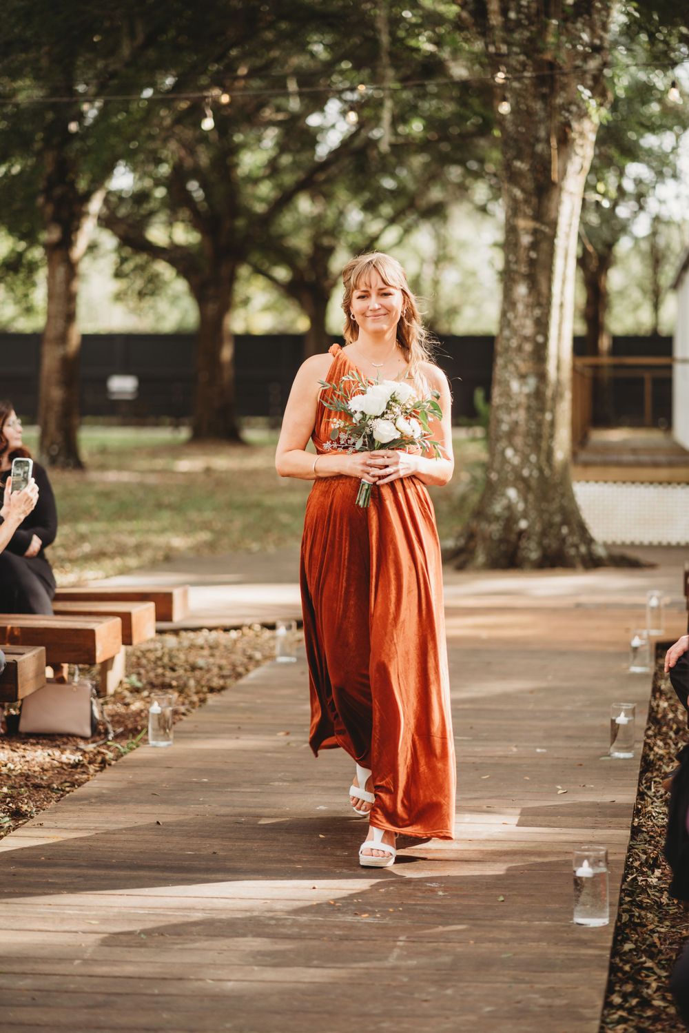 A bridesmaid in a rust-colored dress walks down a wooden aisle carrying a small bouquet.