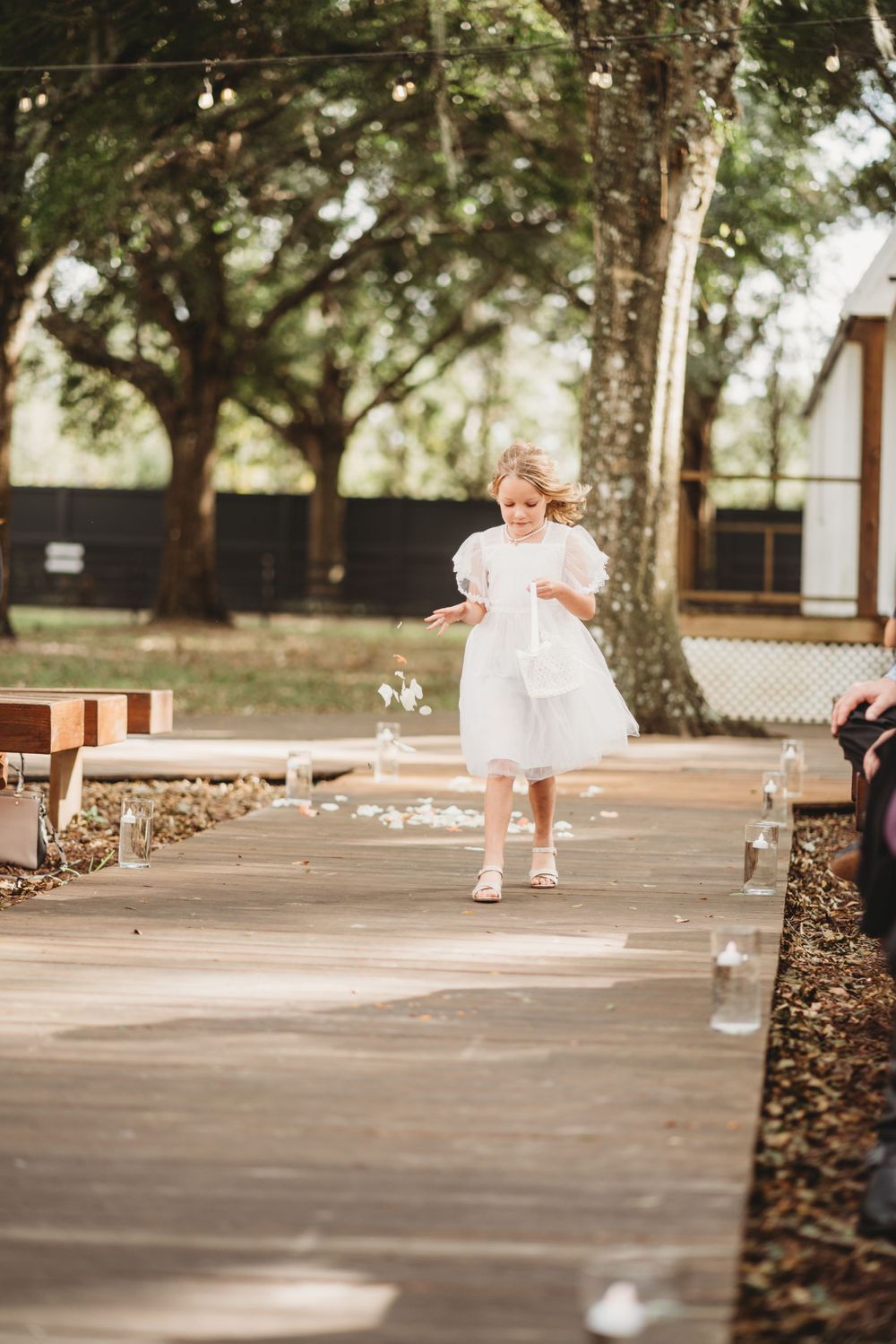 A young flower girl in a white dress walks down a wooden aisle lined with candles.