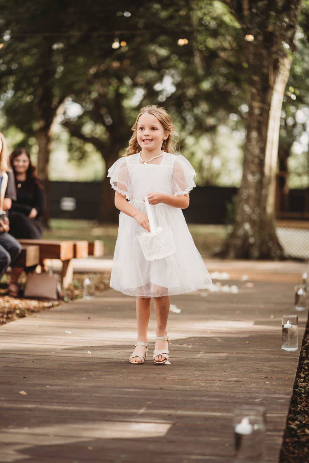 A young flower girl in a white dress with puff sleeves walks down an outdoor wedding aisle.
