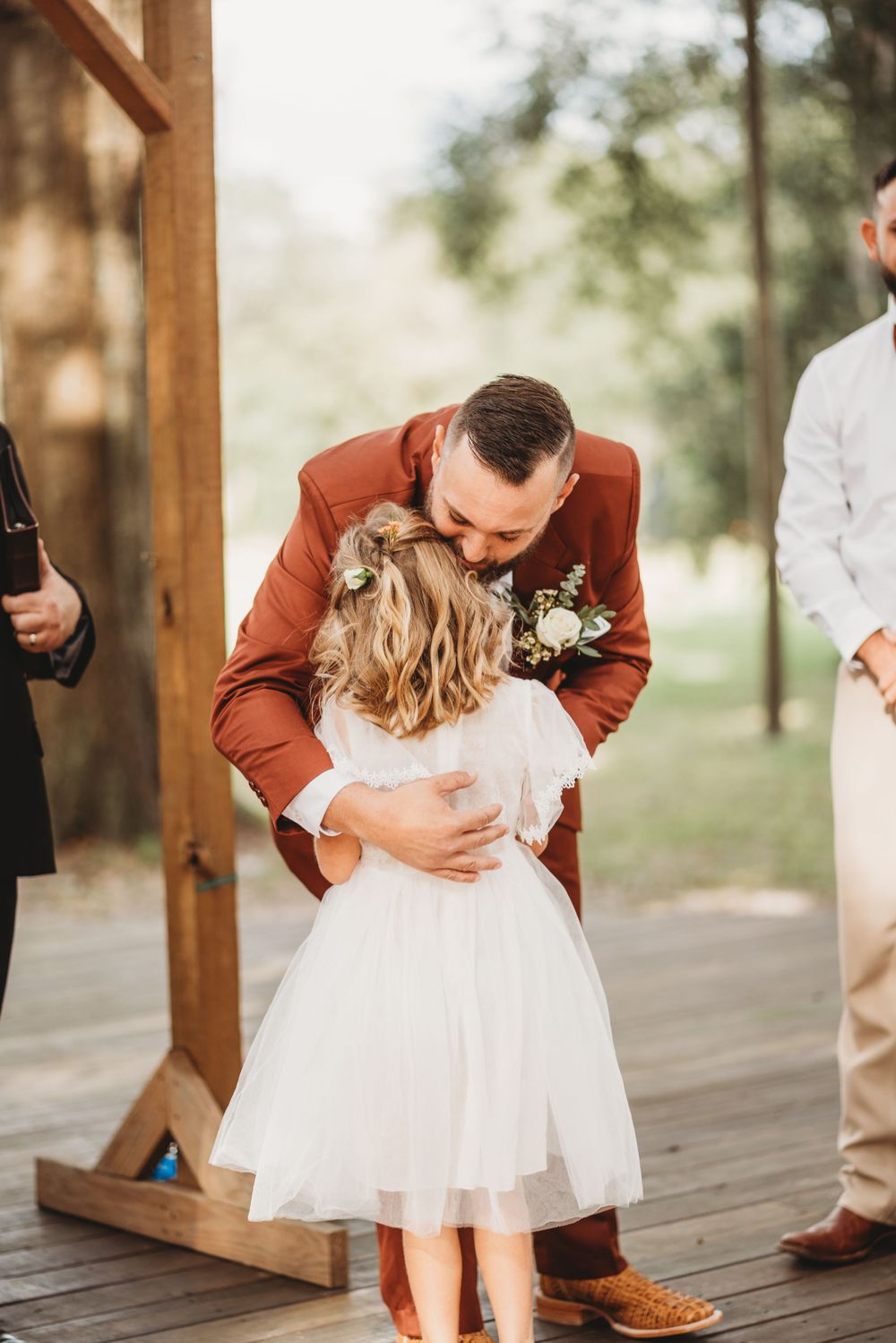 Tender moment between groom in rust-colored suit and young flower girl in white dress at outdoor wedding ceremony.