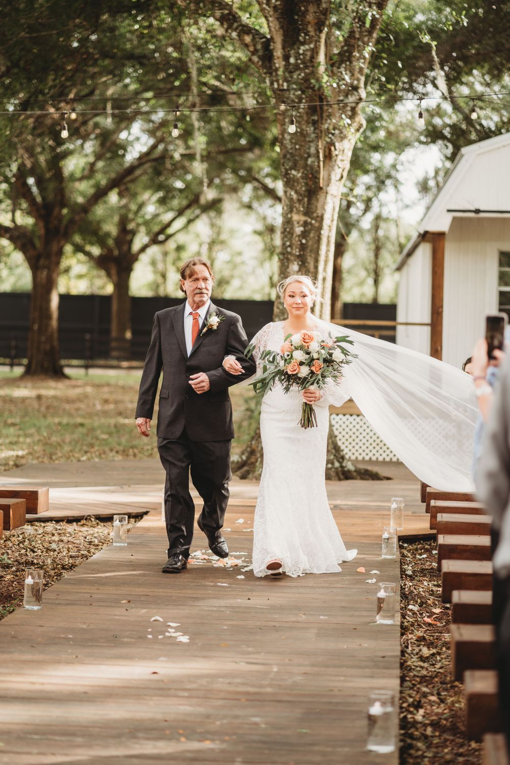 Bride in white lace gown walks down wooden aisle with escort at outdoor rustic wedding venue.