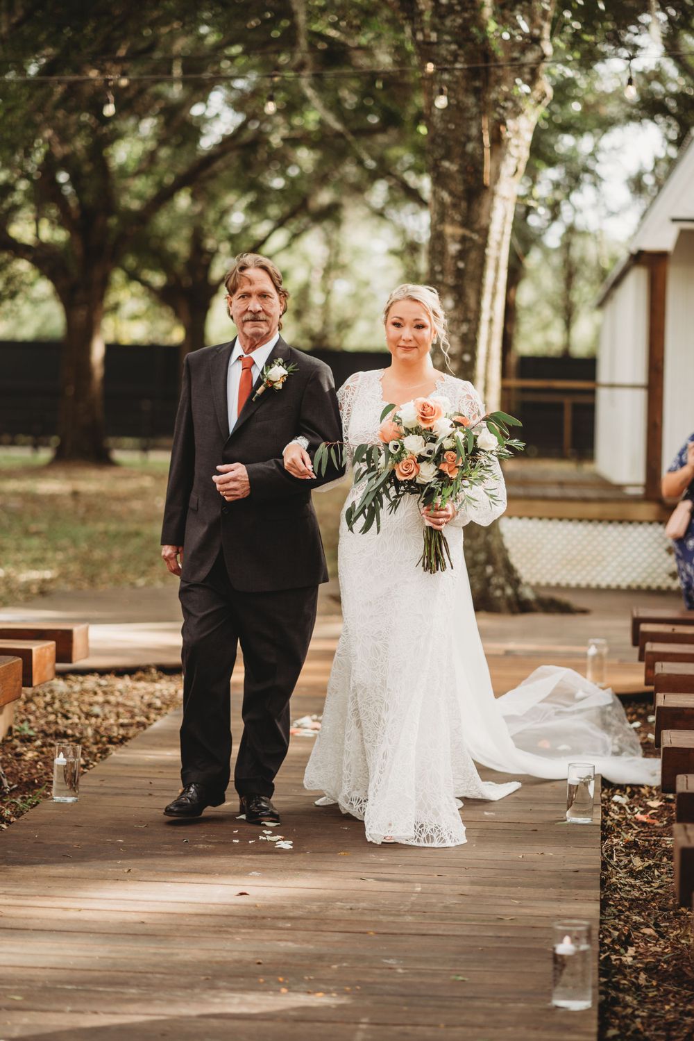 Bride carrying floral bouquet walks down wooden aisle with escort during outdoor ceremony.