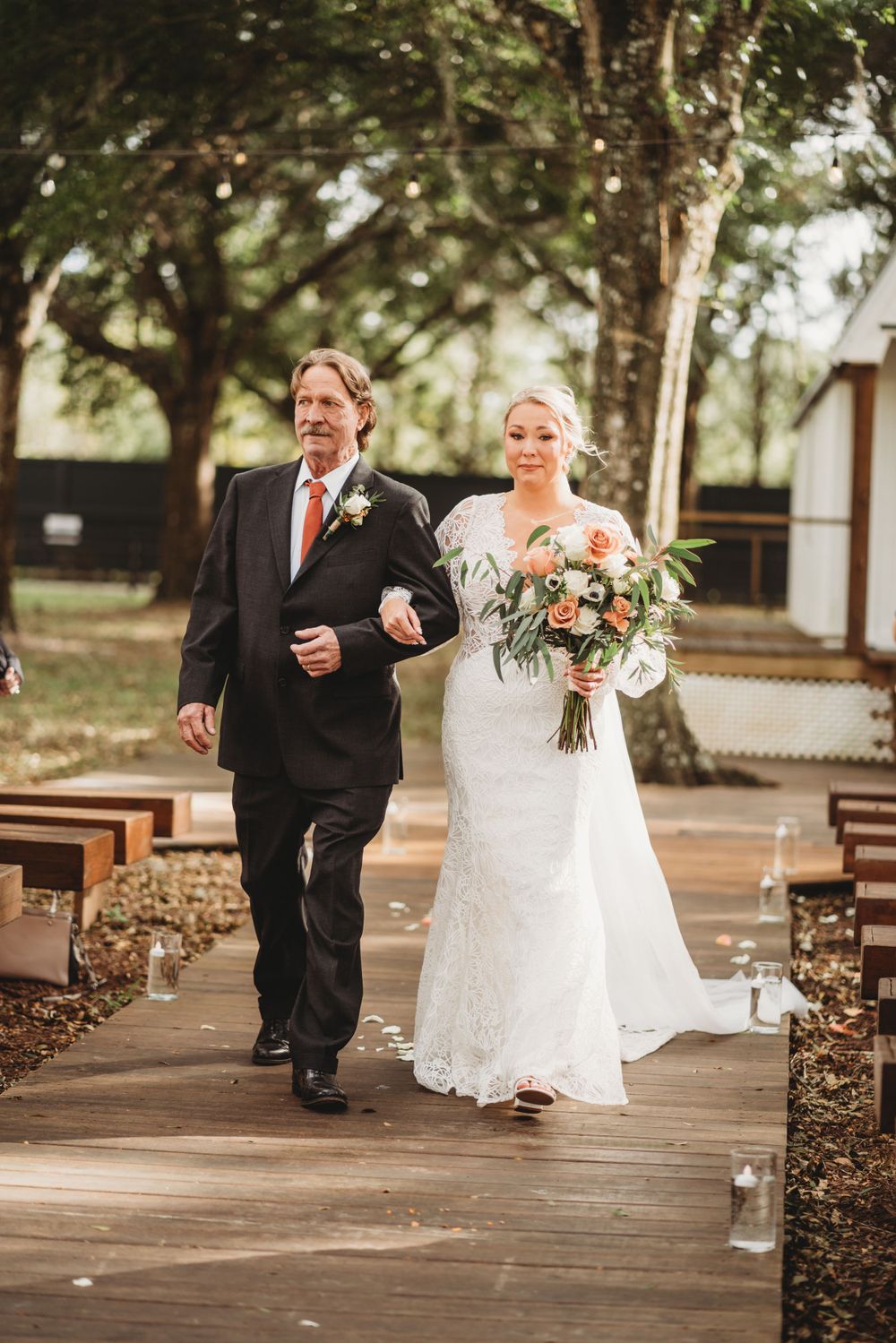 Bride in elegant lace gown walks down wooden aisle with escort at outdoor wedding venue.