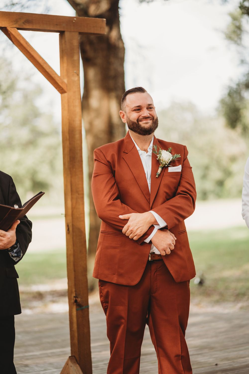 Groom in rust-colored suit stands smiling at wooden altar during outdoor wedding ceremony.