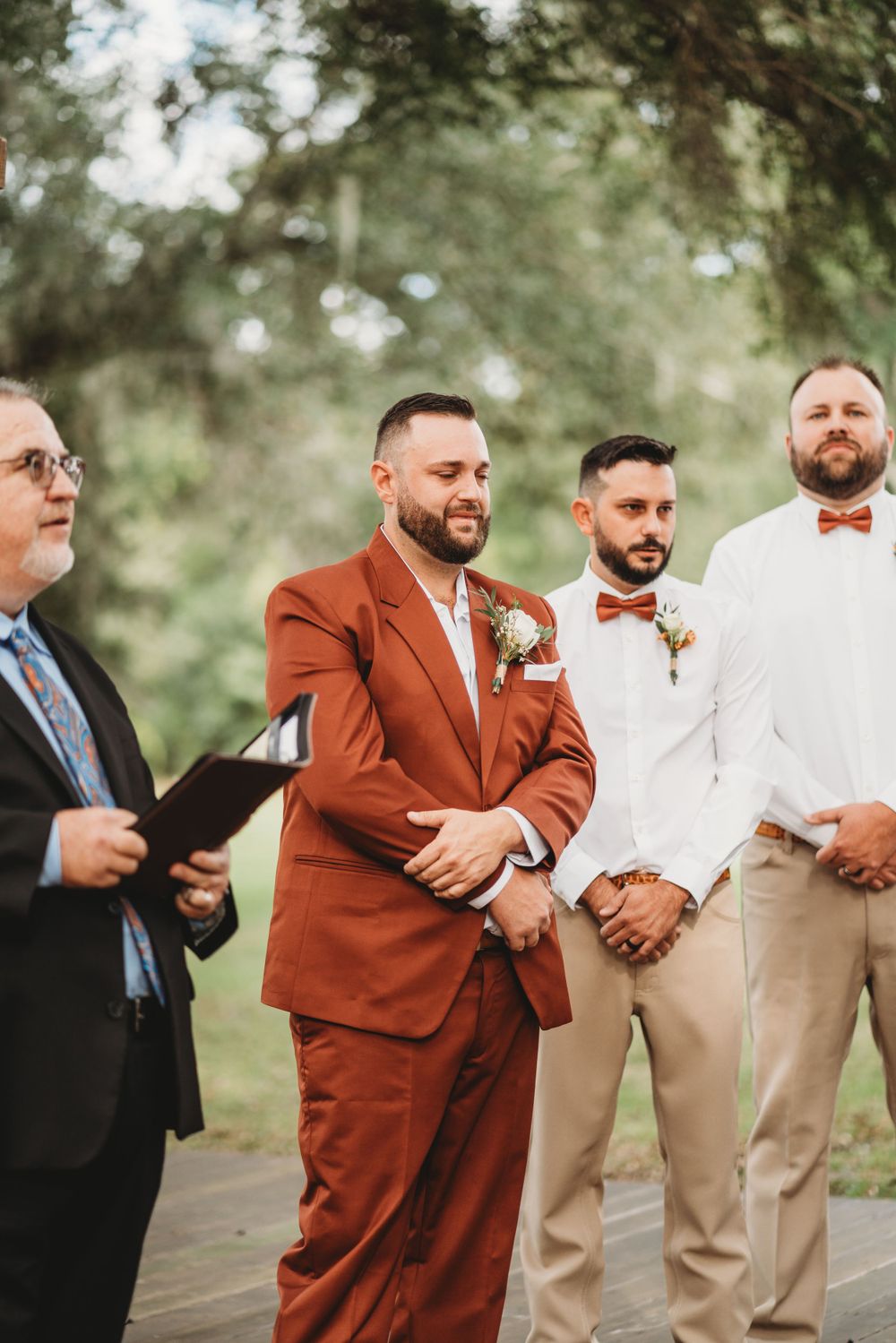 Wedding party stands at rustic outdoor altar with groom in rust suit and groomsmen in light attire.