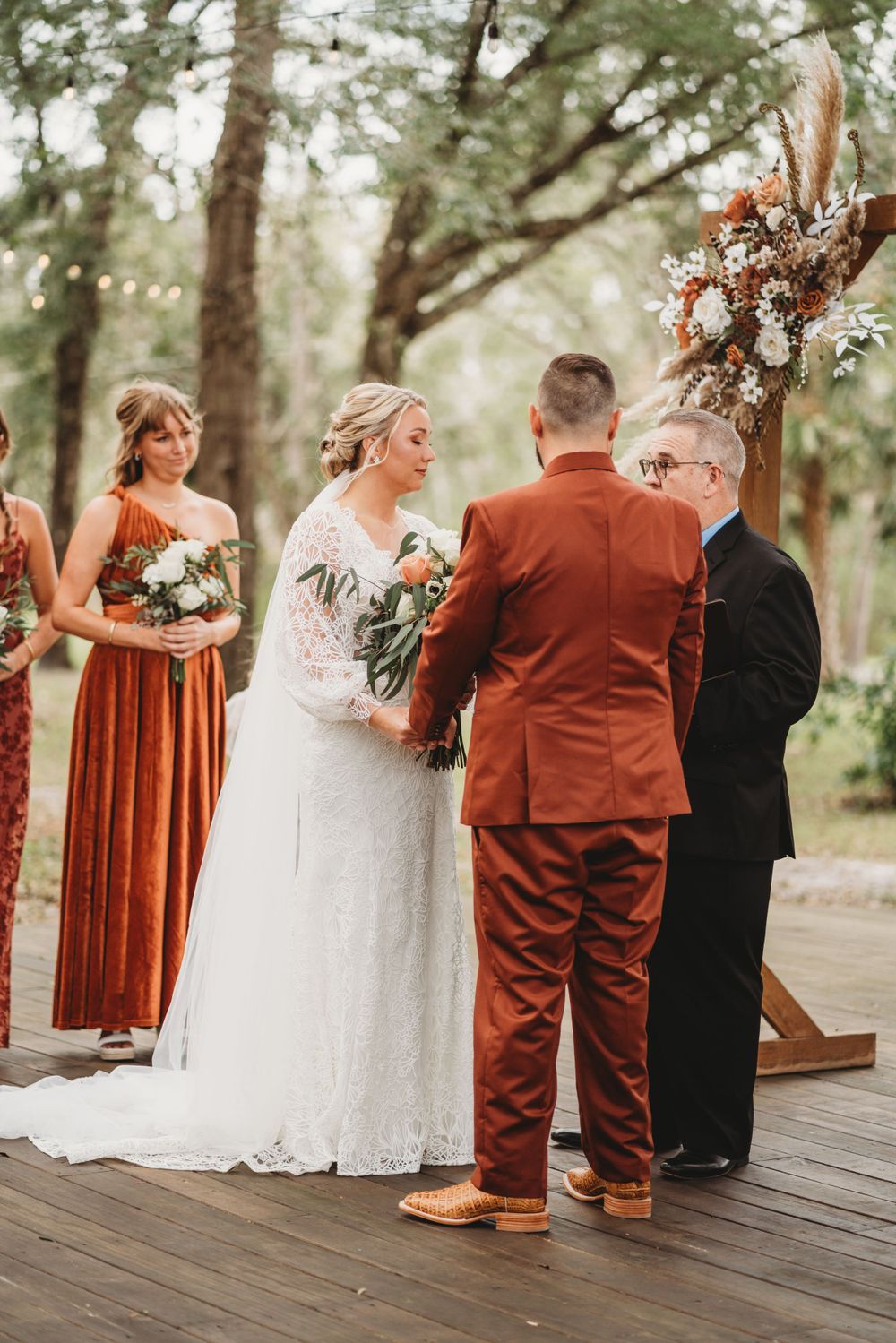 Outdoor wedding ceremony with bridal party in rust-colored attire and wooden arch with floral decorations.