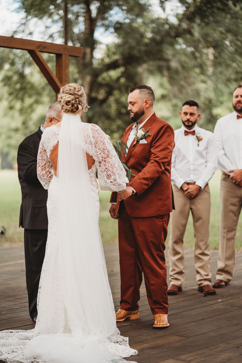Wedding ceremony at wooden altar with groom in rust suit and groomsmen in neutral colors.