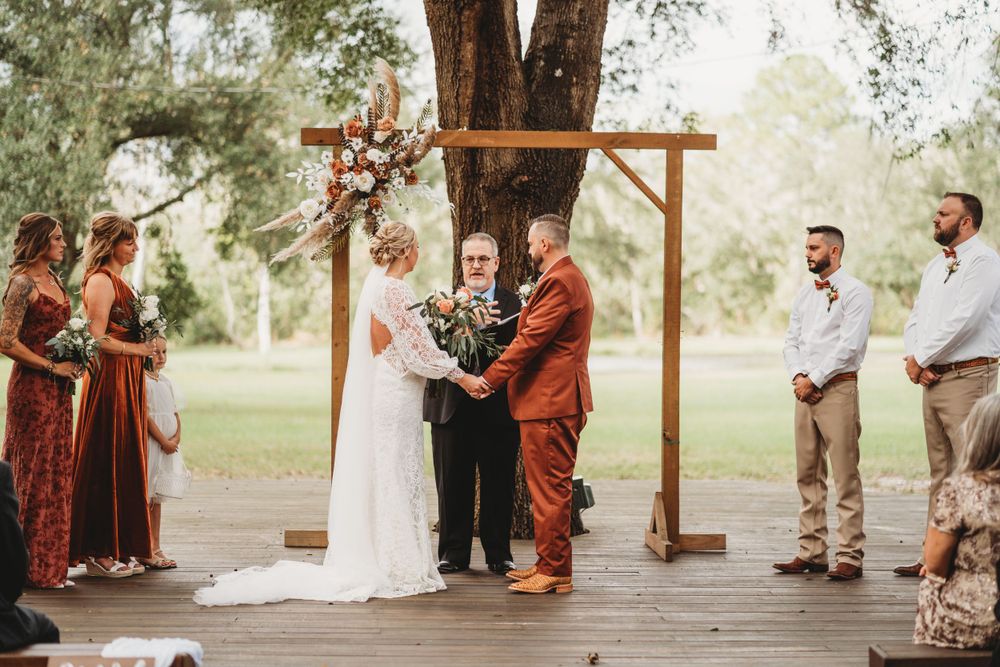 Rustic outdoor wedding ceremony under wooden arch with bridal party in rust and neutral colors.