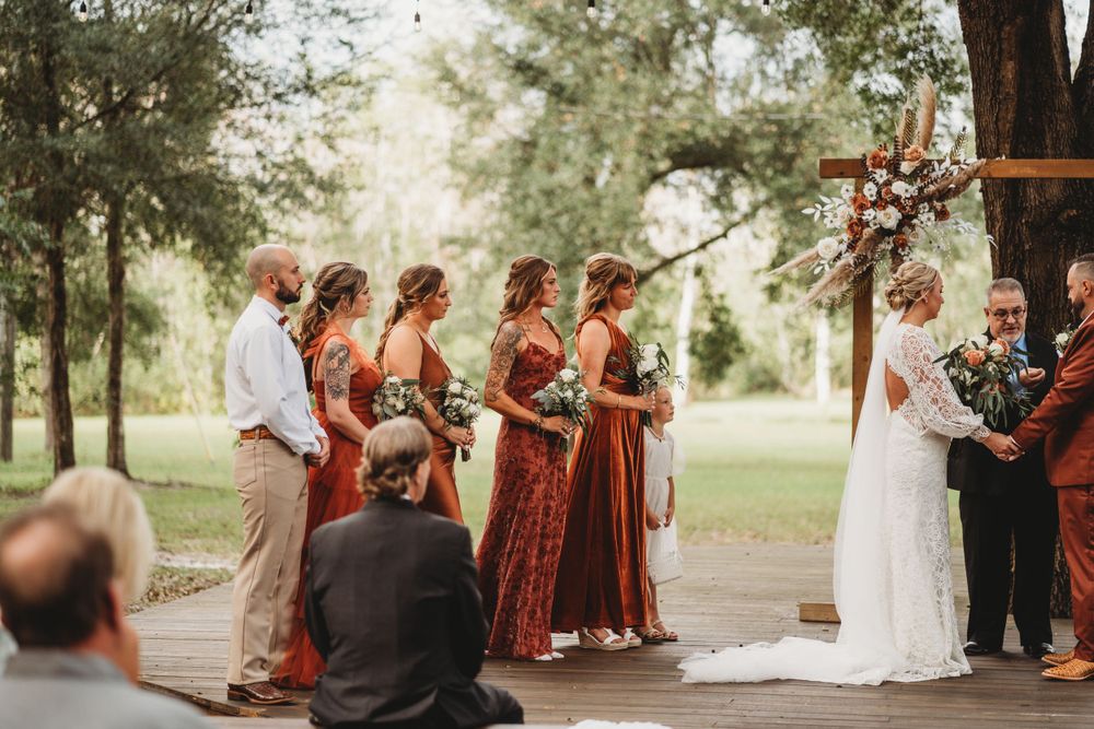 Wedding ceremony with bridesmaids in rust dresses and wooden arch decorated with flowers.